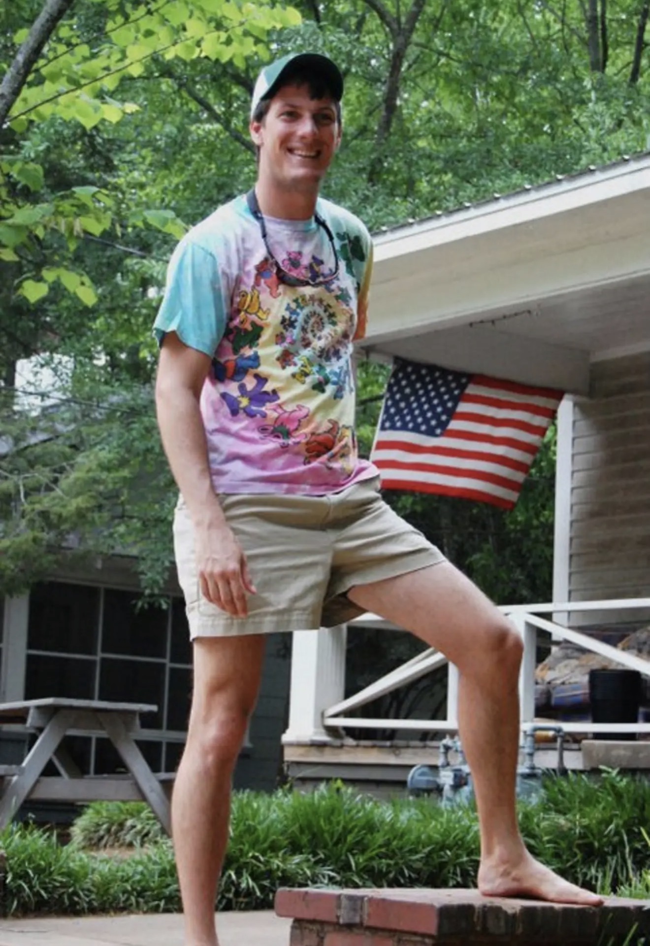 A guy in a tie-dye shirt standing on a brick with the American flag on the porch behind him